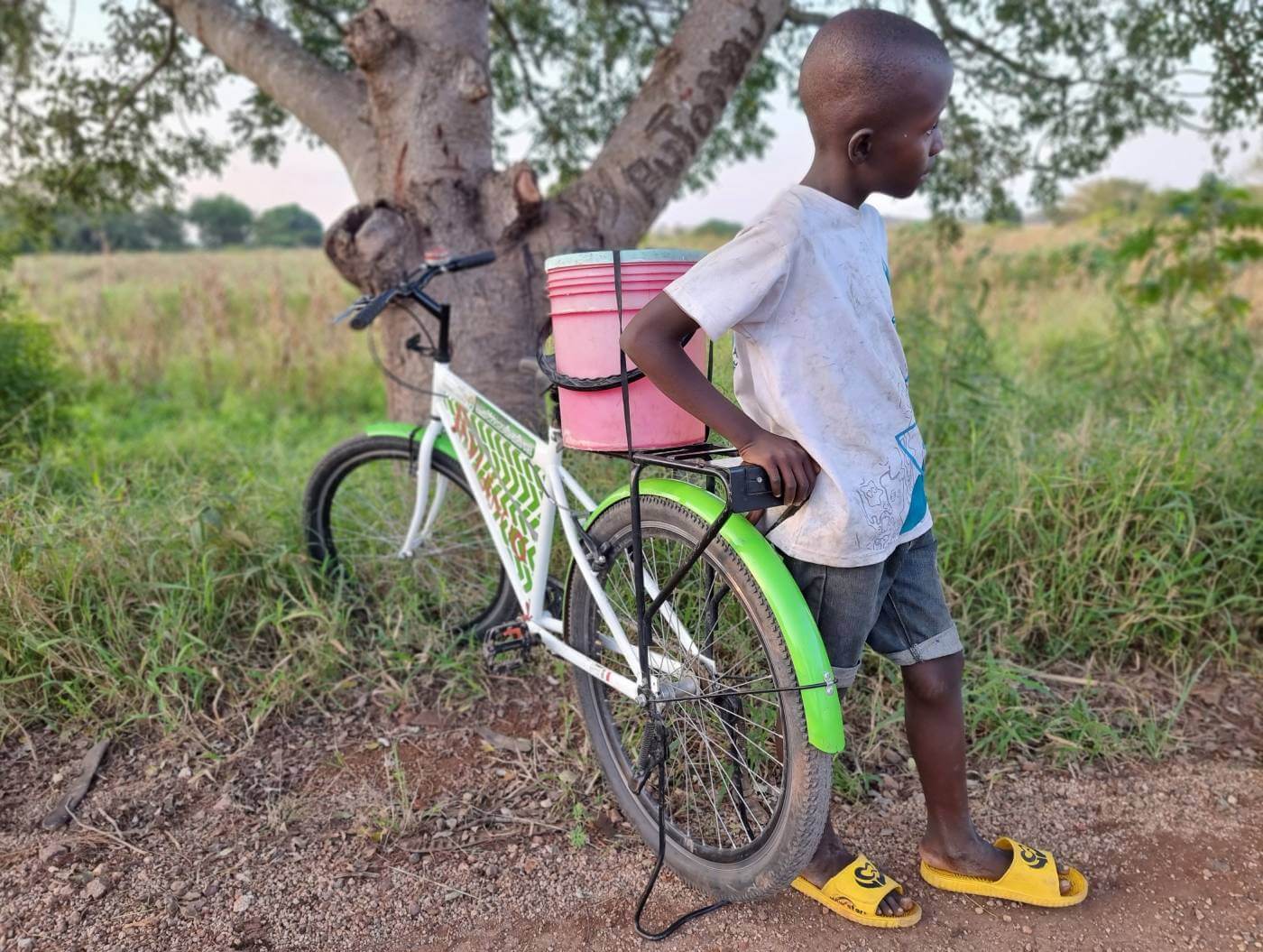 A child stands next to a green and white Mozambike bicycle with a pink and white striped basket on the back, leaning against a tree trunk in a rural setting with a dirt road and green vegetation in the background.