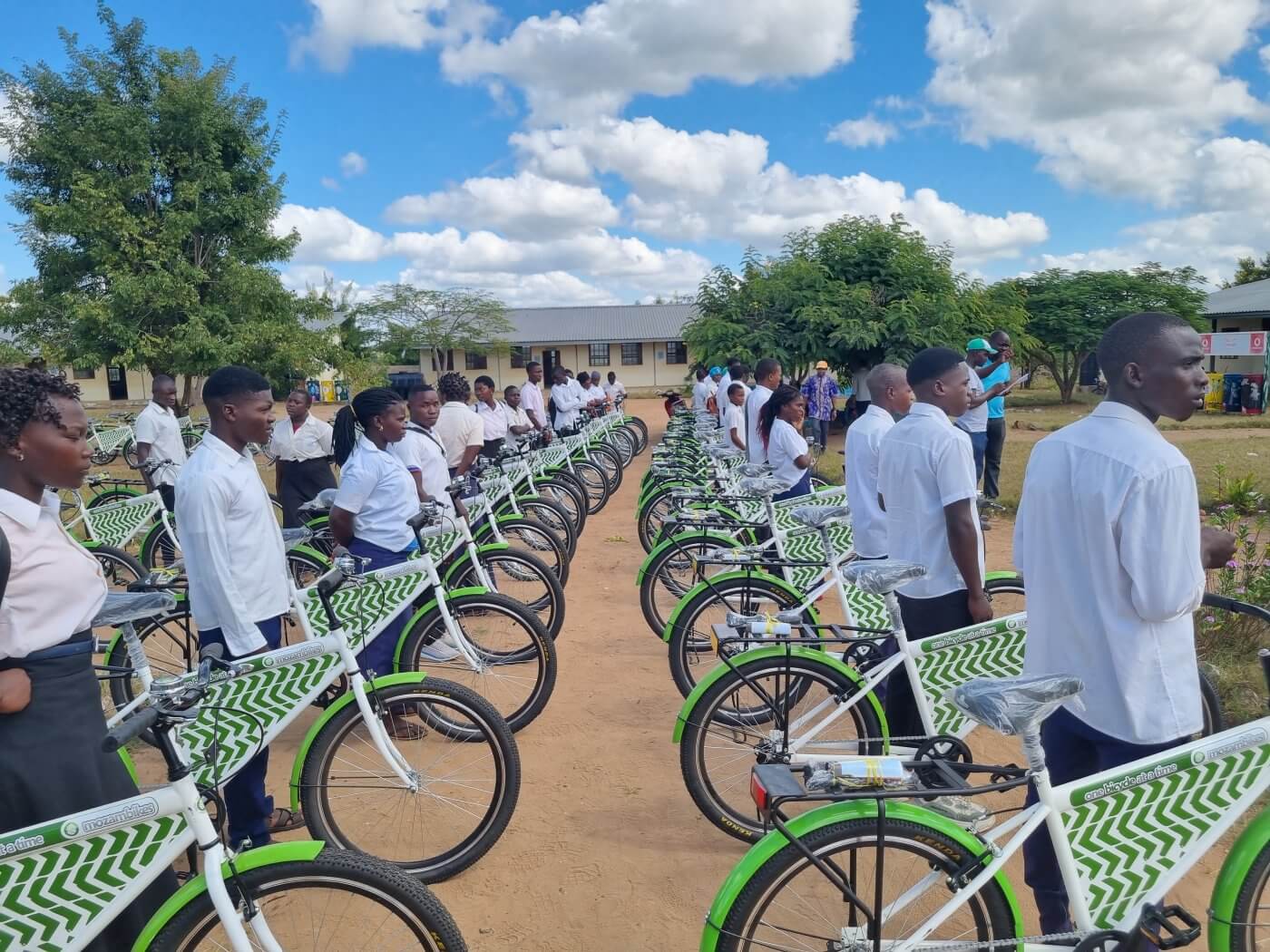 A group of young students wearing white shirts and black pants stand next to a line of green and white Mozambikes bicycles with baskets on the front, parked on a dirt road with trees and a single-story building with a red roof in the background. The sky is blue with white clouds.