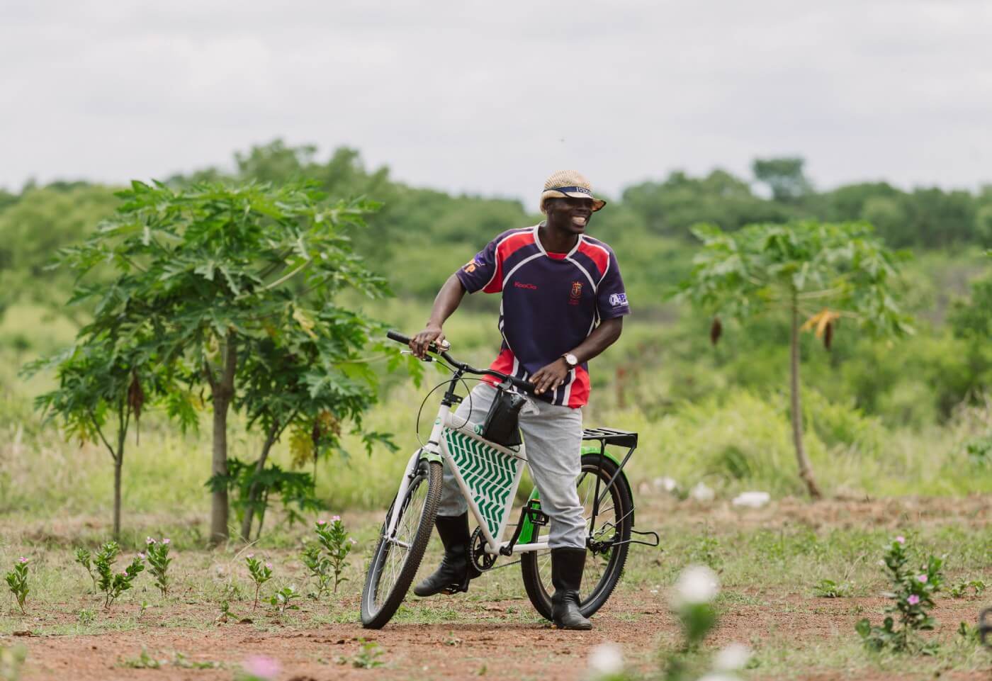 A photo-realistic image of a person wearing a blue and red jersey with a white stripe on the sleeves and a white baseball cap, riding on a green and white Mozambikes bicycle on a dirt path in a rural setting. The path is surrounded by small plants and trees, with a cloudy sky and tree line in the distance.