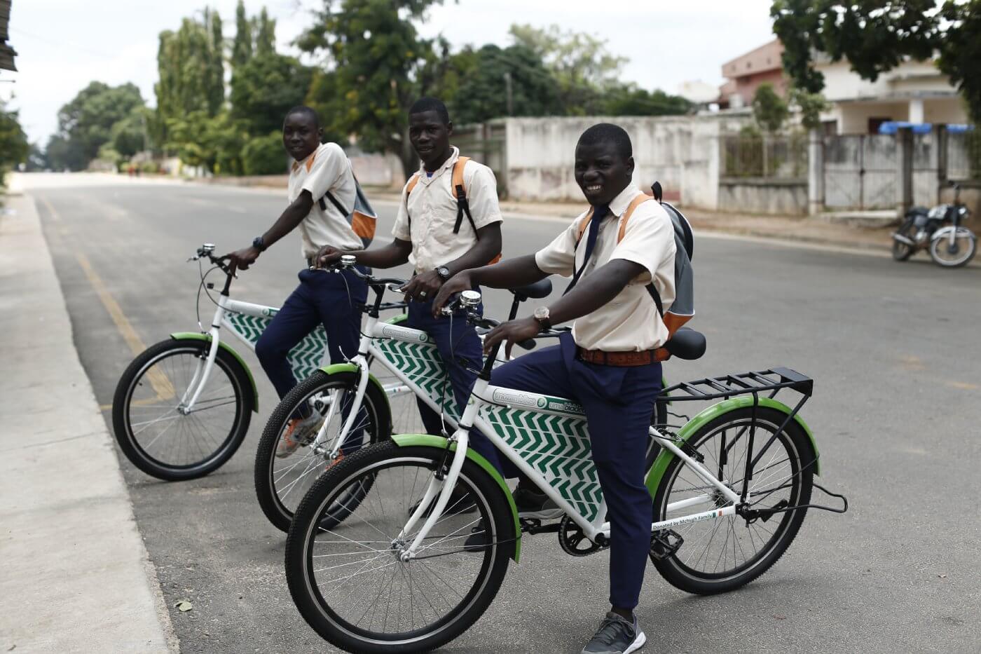 Three people wearing orange shirts and black pants ride green and white Mozambike bicycles with baskets on the back, on a street surrounded by trees and buildings.