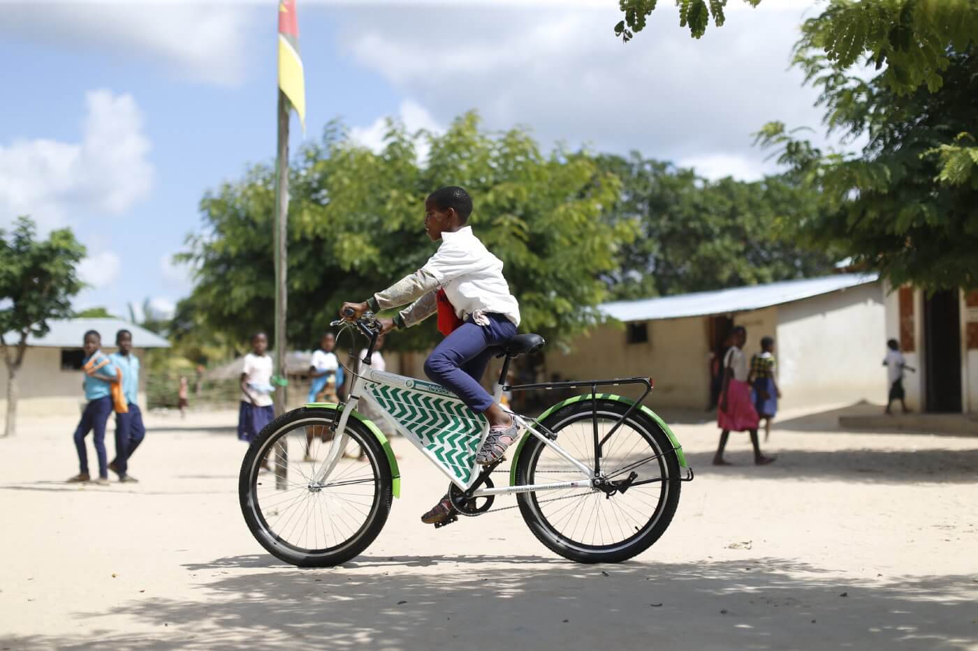 A young boy wearing a white shirt and blue pants patterned school uniform rides a white and green patterned Mozambikes bicycle in a rural school with trees, buildings, and a yellow and red flag in the background.