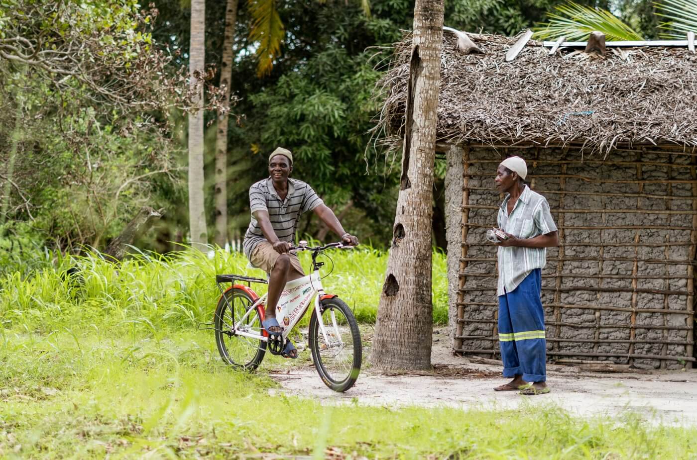 A tropical scene with a man riding a red and white mozambike bicycle and another man standing next to a bamboo hut with a thatched roof. Both men are wearing striped shirts and the background is filled with lush greenery.