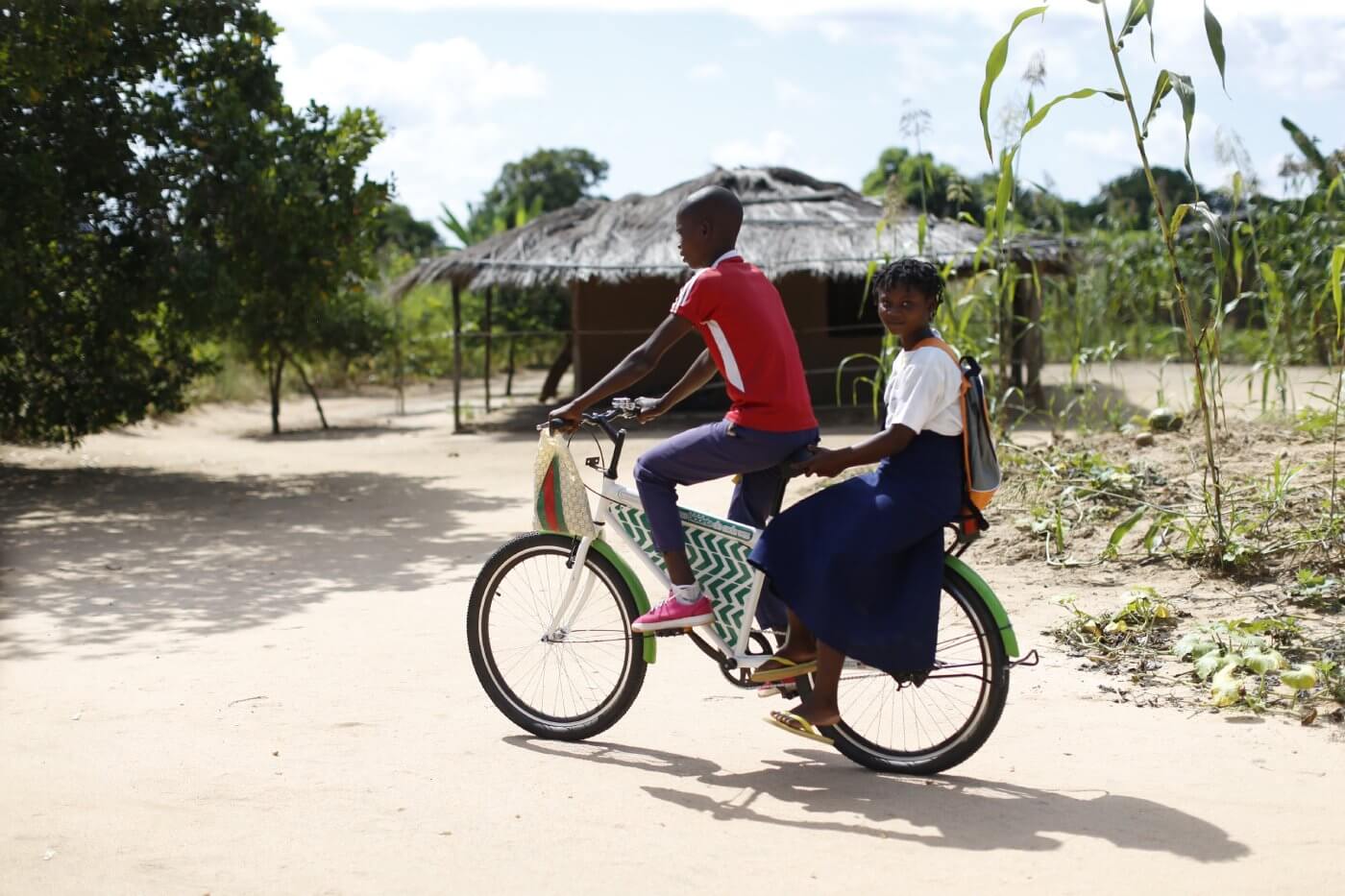 A photo-realistic image of two boys wearing red and blue t-shirts, riding on a green Mozambikes bicycle with a white frame and green handlebars on a dirt road in a rural setting. In the background, there is a thatched roof hut and trees, with a blue sky and white clouds above.