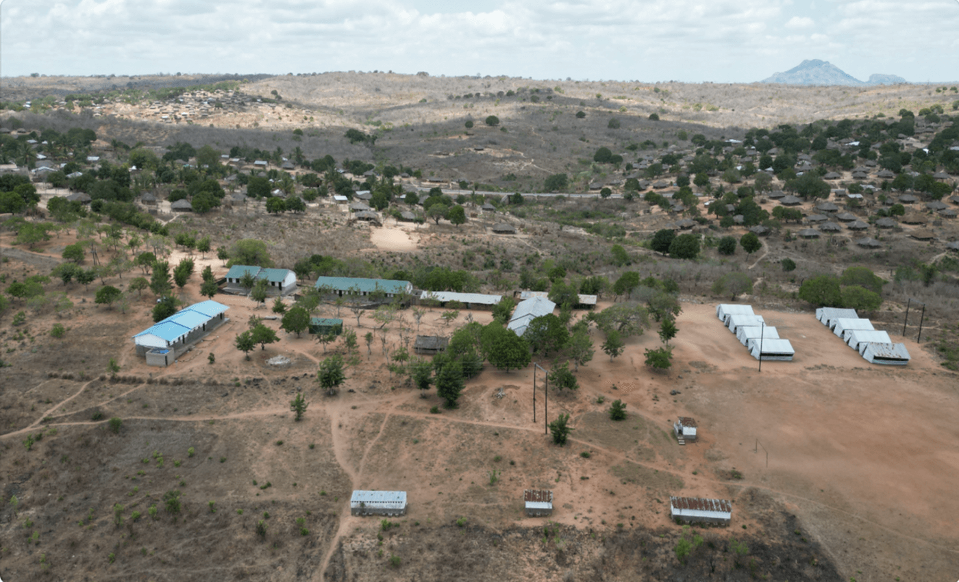 This photo shows an aerial view of a school in a rural village in Africa. The school is made up of small houses with tin roofs and a few larger buildings with blue roofs. The village is surrounded by dry shrubland and rocky hills, with a few trees scattered throughout. The sky is cloudy, giving the image a desolate mood. Despite the challenging environment, the school stands as a beacon of hope and education for the local community