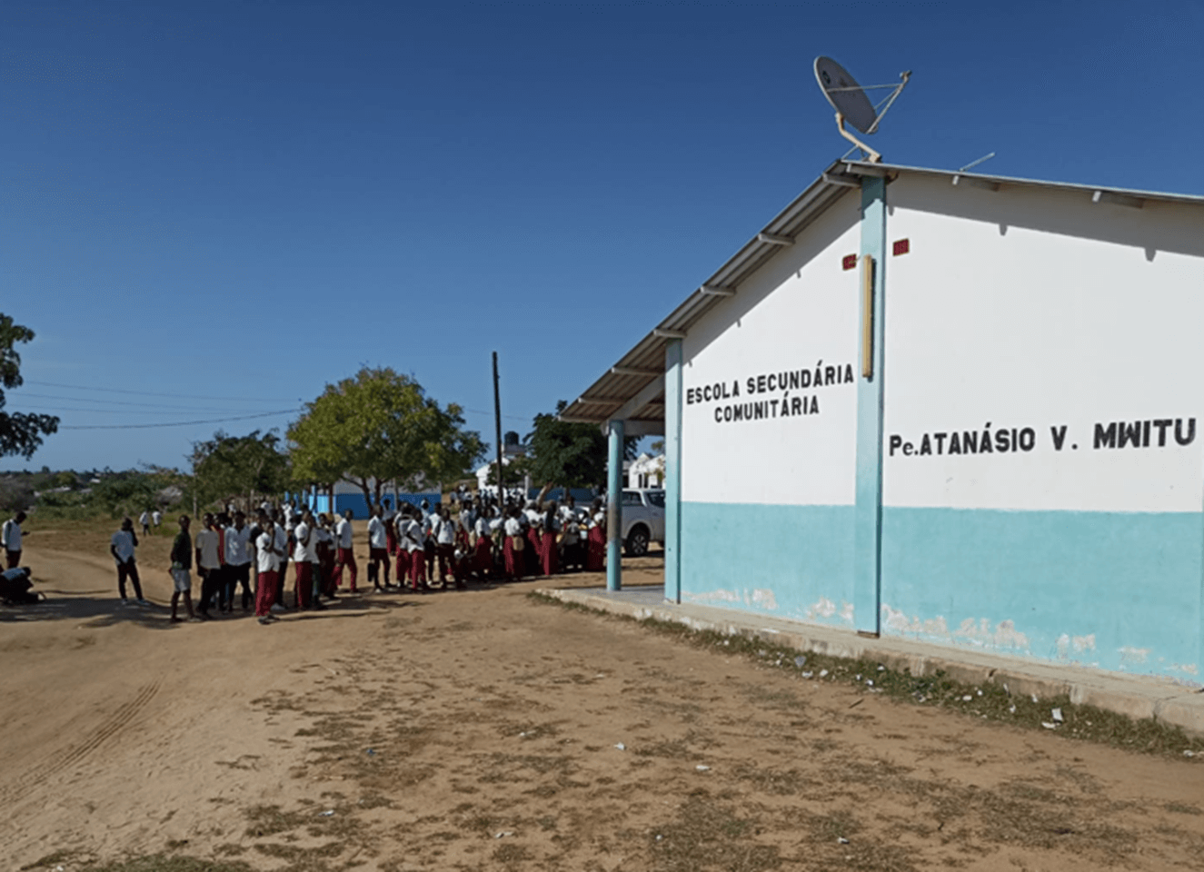 This photo captures a moment at the “Escola Secundária Comunitária Pe. Atanásio V. Minitu” in Mozambique. A group of students in red and white uniforms are gathered outside the white and blue trimmed school building, which has a satellite dish on its roof. The sky is blue and trees can be seen in the background, while the ground is dirt and a dirt road runs in front of the building. This image portrays a vibrant school community in a rural setting.